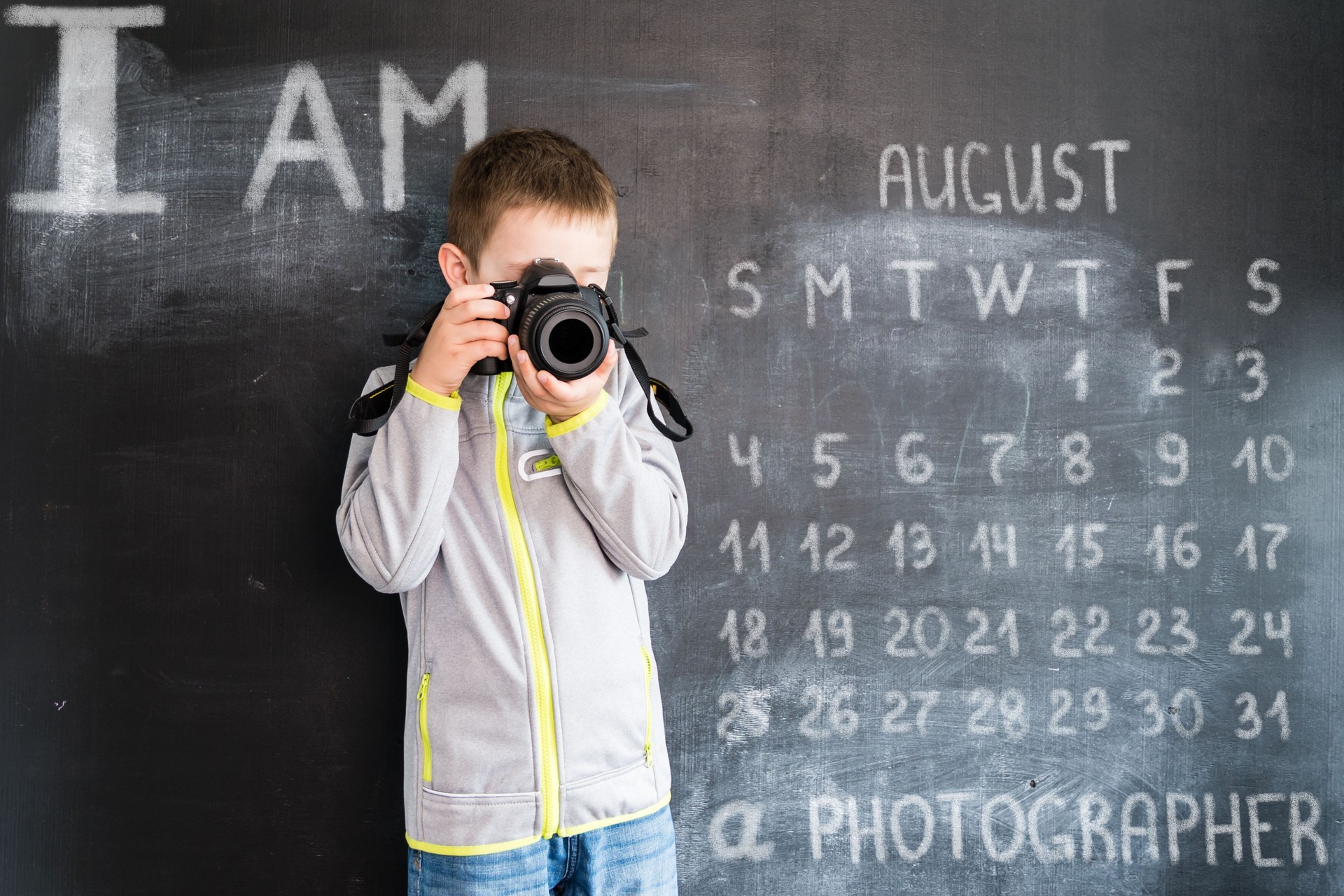 Kleiner Junge ist mit Foto-Kamera in der Nähe von Blackboard schießen. Junge Fotografen. Kreative Design-Konzept für 2019 Kalender. August